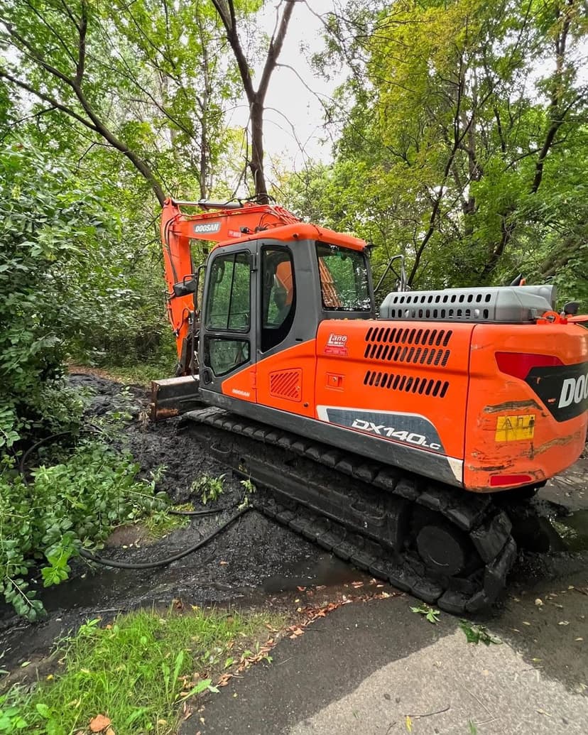 Orange excavator working on a muddy path surrounded by trees and greenery.