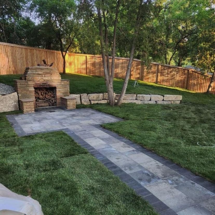 Outdoor landscaping featuring a stone fire pit, paved path, and lush green grass in a fenced yard.